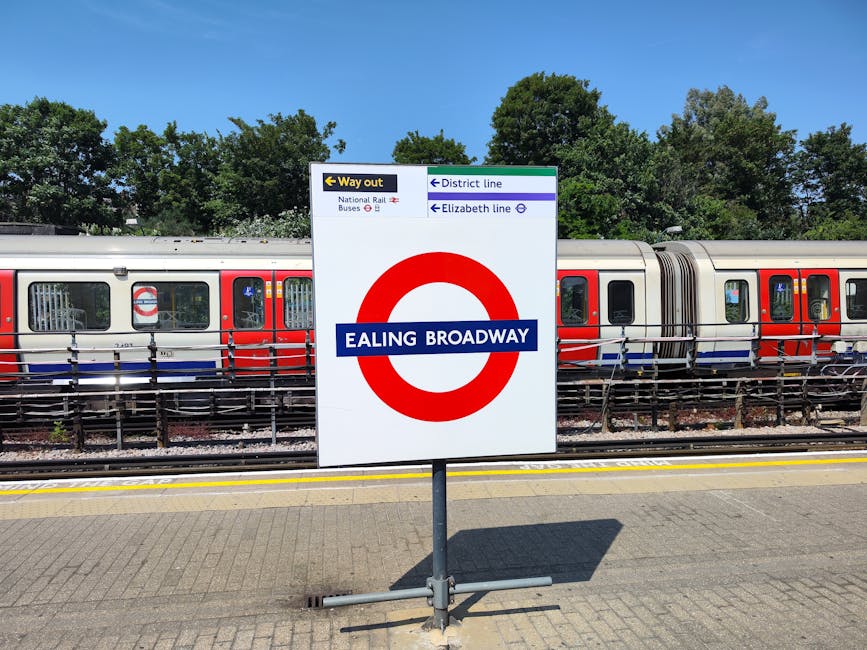 A large rectangular white sign with a red circular London Underground symbol and blue stripe across the middle displaying 'EALING BROADWAY' in white text is positioned on a paved platform at Ealing Broadway station. The sign provides directions for the station's district and Elizabeth line, with additional signage indicating the way out via the national rail buses. Behind the sign, a modern London Underground train with red, white, and blue livery is stationary on the tracks, with visible windows, doors, and safety bars inside. The train is situated behind a metal fencing barrier. In the background, there are tall green trees and a clear blue sky, indicating a sunny day. This scene captures the typical setting of a London transport interchange, useful for illustrating station access in the context of house removals or relocations involving transport logistics managed by Removal Company Ealing.
