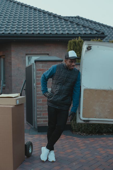 A man dressed in a black padded vest, blue long-sleeve shirt, black trousers, and white footwear is standing outdoors beside a white delivery van with its rear doors open during daytime. He is leaning against the van, looking slightly to his left with his right hand on his hip. In front of him, there are several cardboard boxes placed on a trolley, ready for loading or unloading. The scene is set in a residential area with a brick house featuring a tiled roof, a partially open gate, and surrounding greenery. The area is paved with brick, and natural daylight illuminates the scene, reflecting a typical home relocation process involving packing, loading, and transport services from Removal Company Ealing, tailored to challenging access situations like tight driveways or narrow doorways.