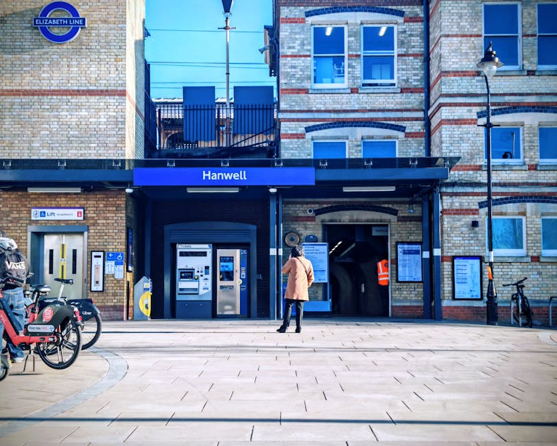 A woman stands outside the entrance of Hanwell Underground Station, part of the Elizabeth Line, on a clear day. The station's brick facade features a blue sign with white lettering displaying 'Hanwell' above the entrance. To the left, there is a row of bicycles including a bike sharing scheme with branded lockers. On the right side, a streetlamp and pavement signs are visible alongside notices posted near the entrance. The entrance itself includes ticket machines, an automated fare collection gate, and an underground passageway leading into the station. The surrounding area has a paved pedestrian walkway, suitable for loading or moving large items, and is part of an urban environment relevant to home relocation or furniture transport planning. The scene captures the typical setting where removal companies, such as Removal Company Ealing, might organize logistics for moving or packing operations, particularly in homes close to public transport links on busy streets.
