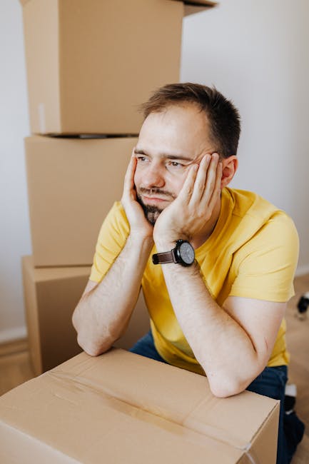 A man with short dark hair and a beard, wearing a yellow t-shirt and a black wristwatch, sits at a table in an indoor space surrounded by packed cardboard boxes. He rests his elbows on the table and s